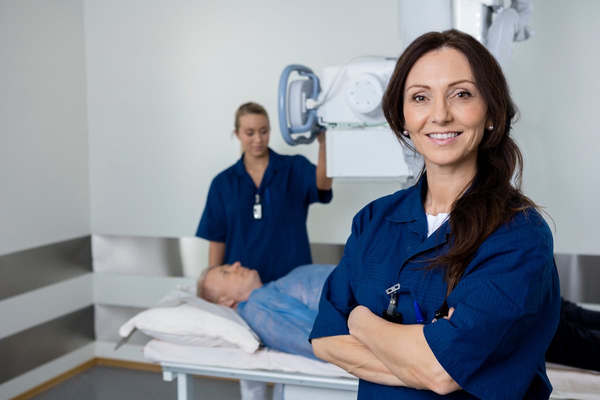 A radiologist smiling a the camera. Behind her, a man lays down on a table and another radiologist sets up an x-ray.