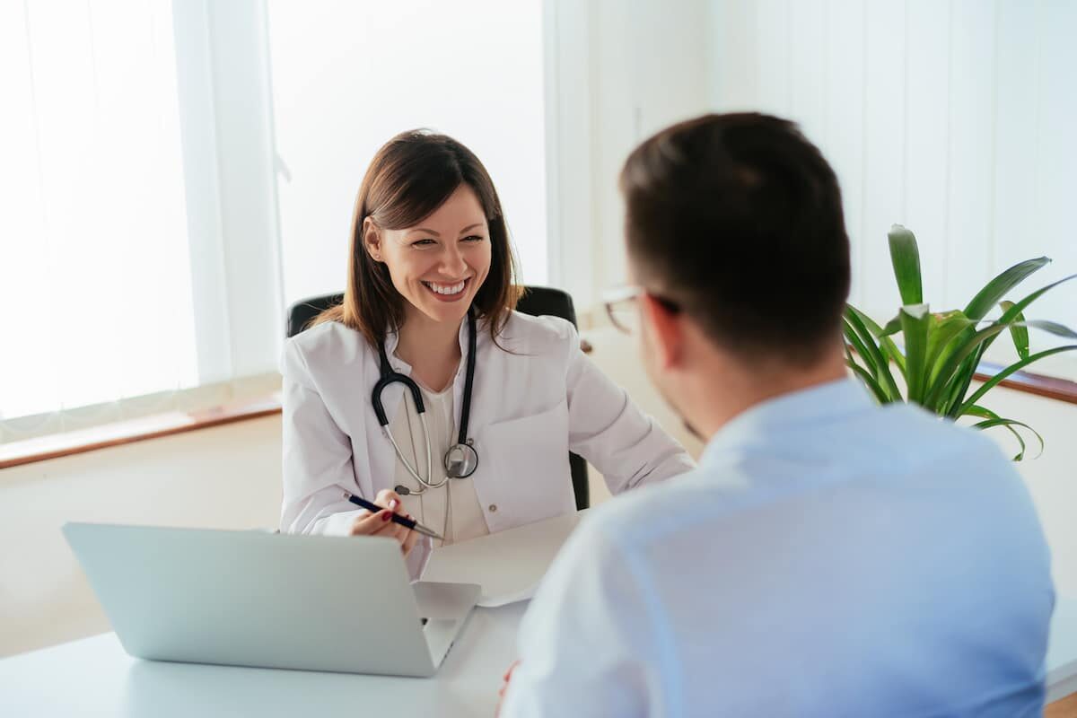 A female doctor sitting at her desk with her laptop and clipboard, talking to a colleague.