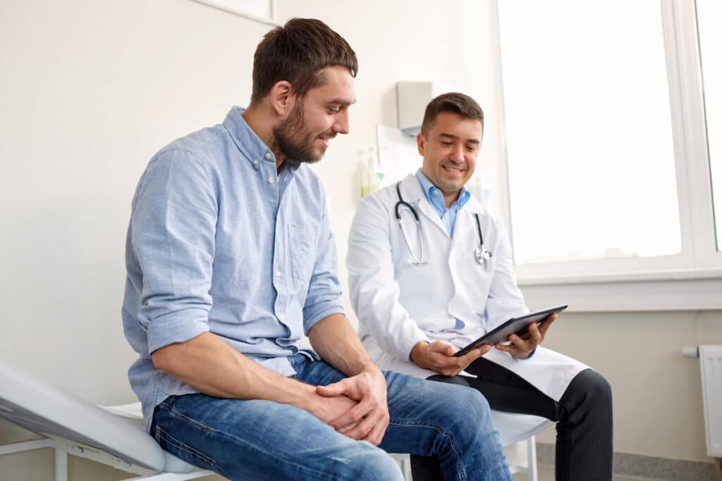 A doctor sitting on a lab table with his patient, showing him notes on the tablet.