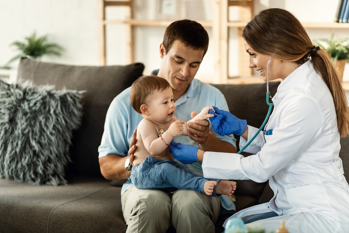 A pediatrician checking a baby's chest with a stethoscope as his dad holds him on his lap.