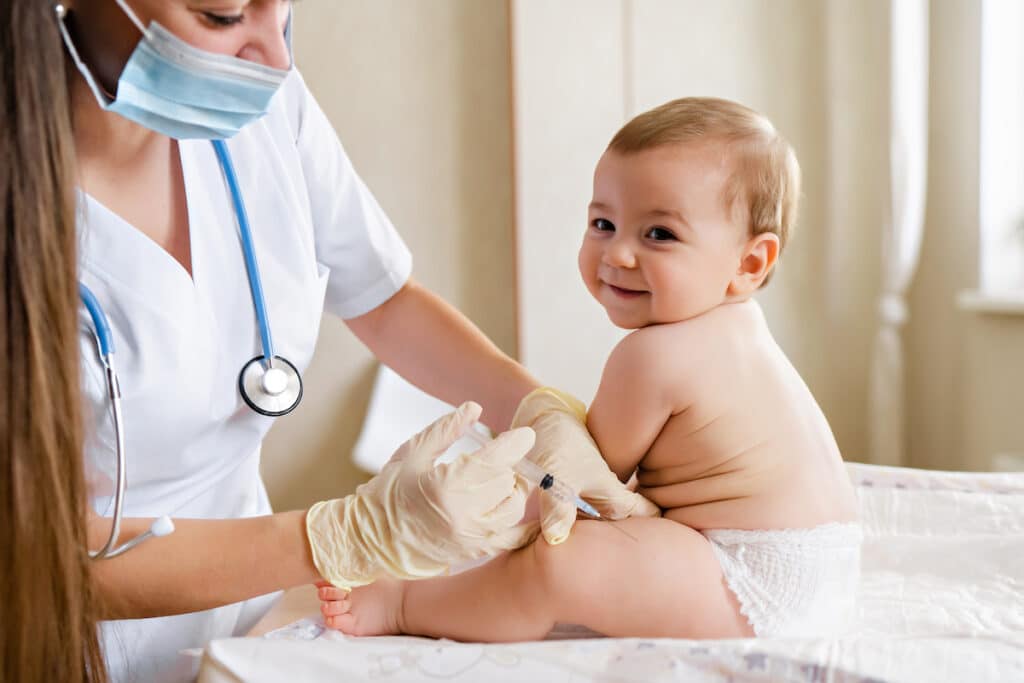 A baby sitting on an exam room table, smiling as a nurse goes to give him a shot in his thigh.