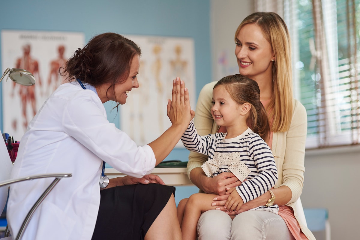 A pediatrician high fiving a young girl as she sits on her mom's lap after an appointment.