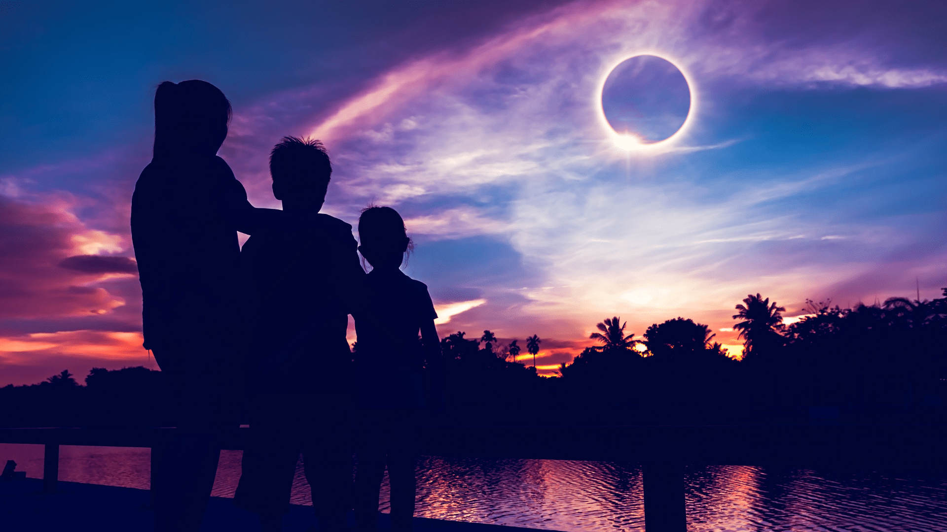 A mom and two kids stand silhouetted by a lake as the total solar eclipse occurs.