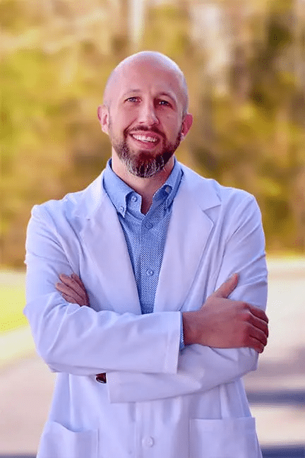Professional headshot of Clark Roberts, APRN, a Healthy Connections team member wearing his medical coat and standing outside with his arms crossed.