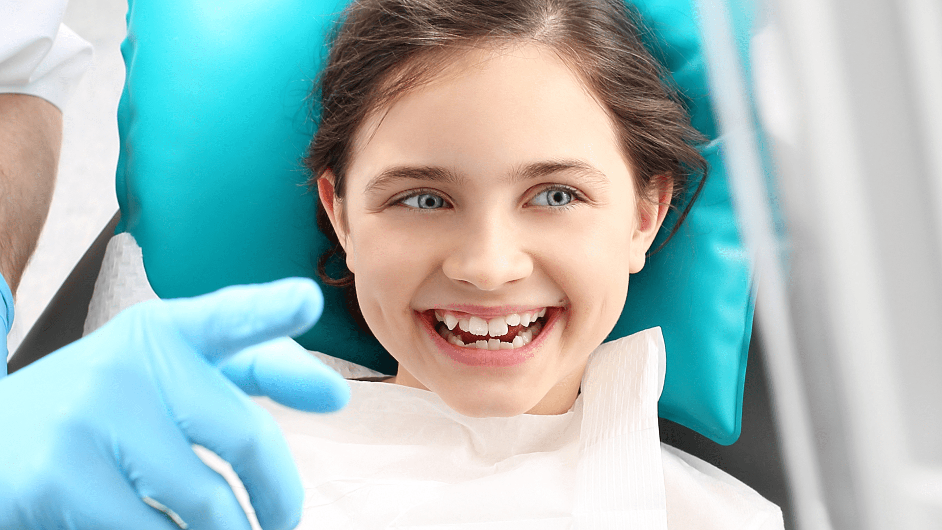 A young girl lays down on a dentist chair, smiling as a dentist points at something over to the side.