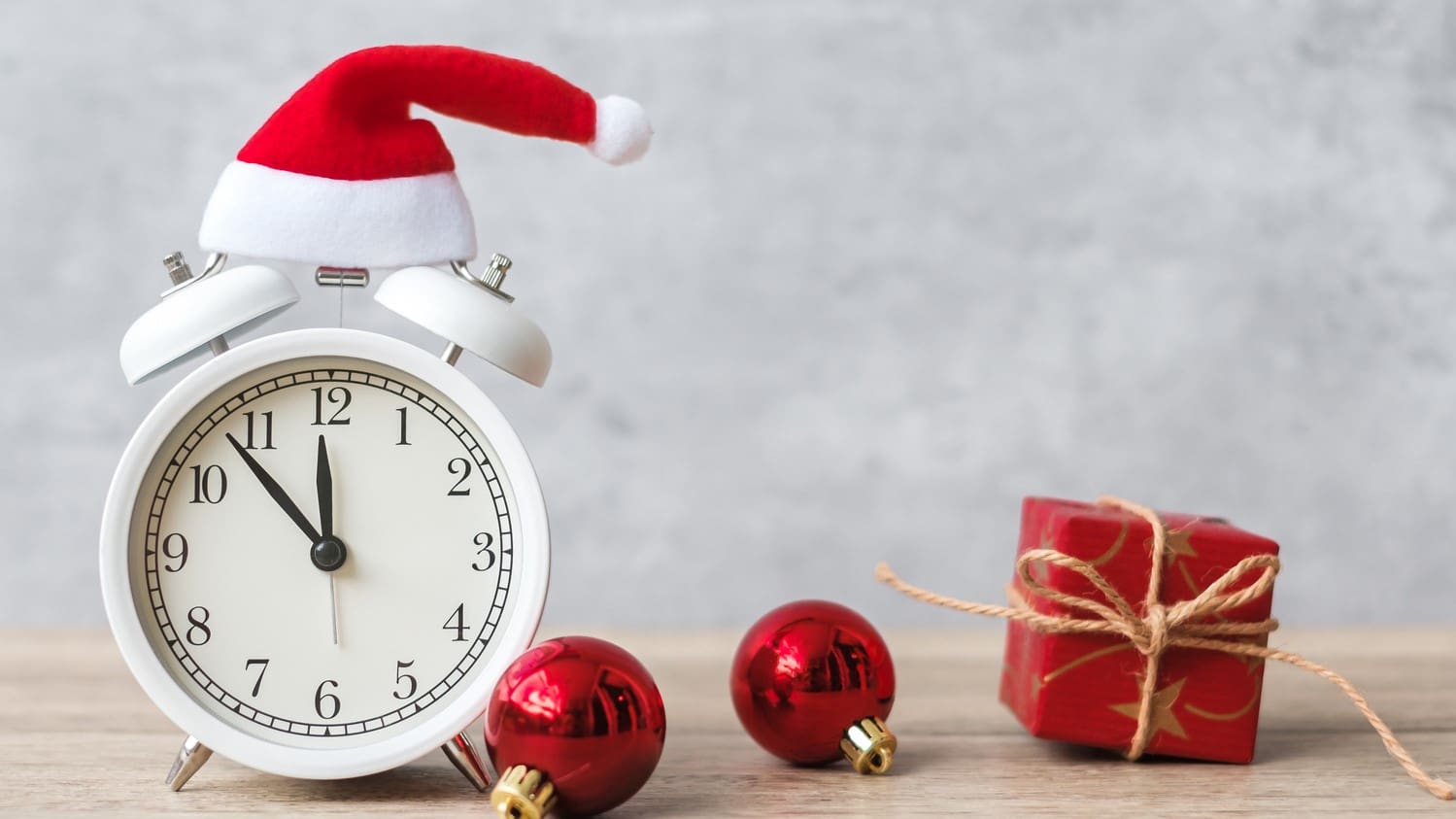 A white alarm clock with a small santa hat on it on a desk next to two red ornaments and a small red wrapped present.