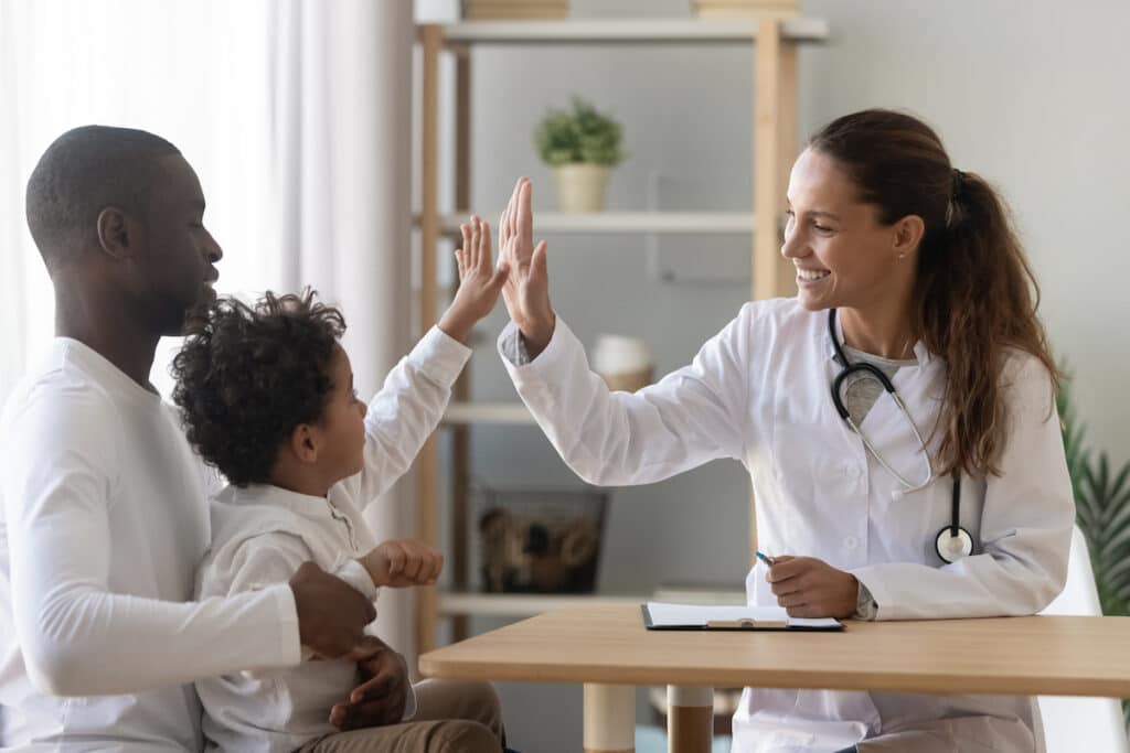 A doctor high fives a young boy as he sits on his dad's lap.
