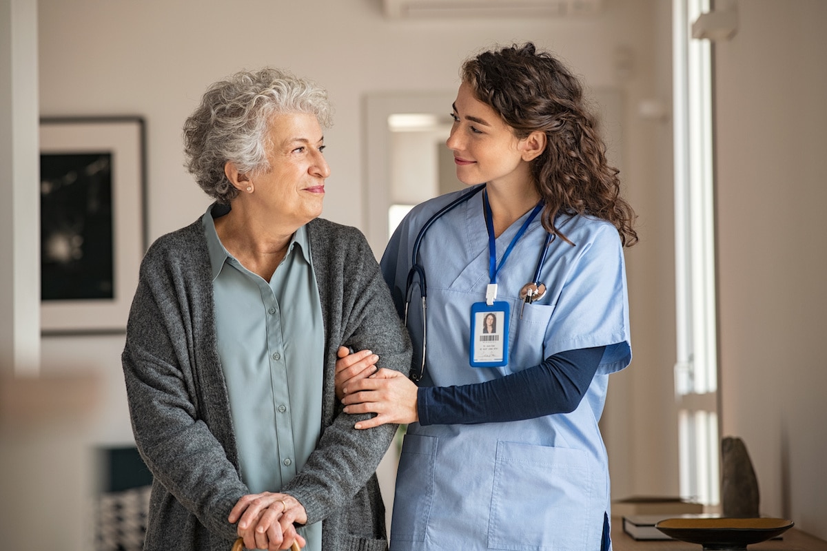 A nurse links arms with an elderly patient, walking down the hall.