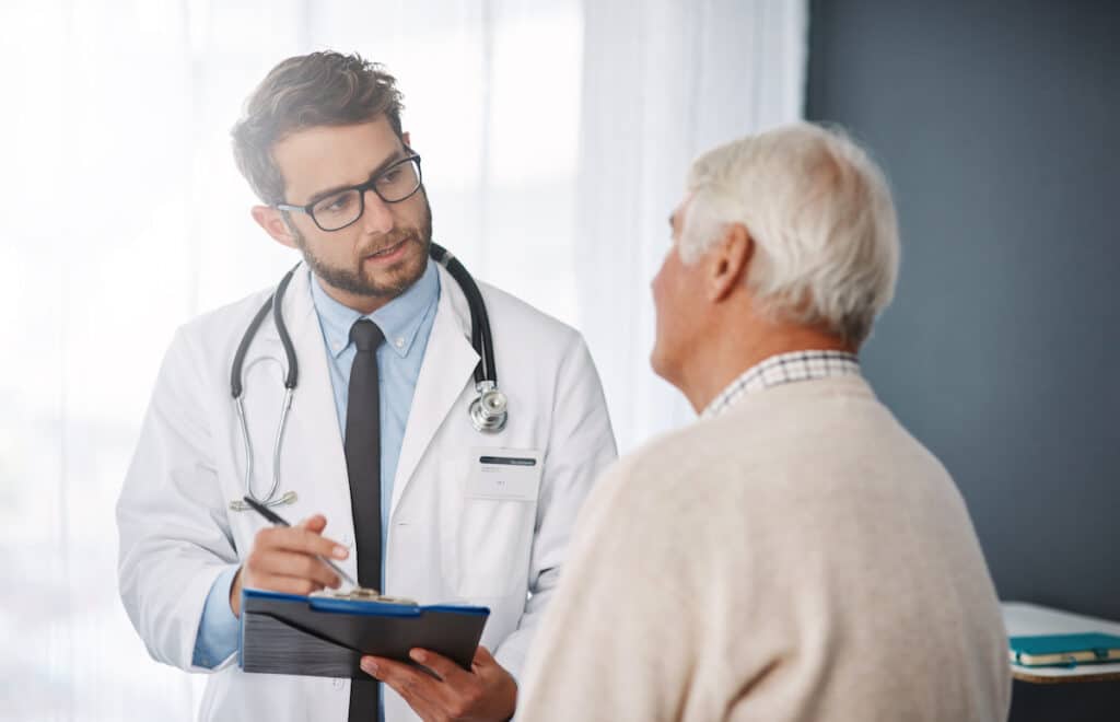 A doctor making notes on a clipboard as he talks to an elderly patient.