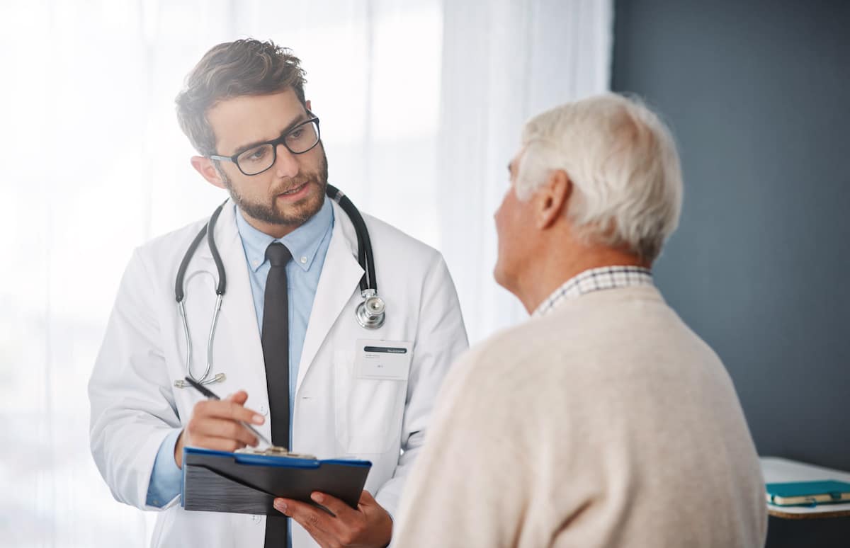 A doctor making notes on a clipboard as he talks to an elderly patient.