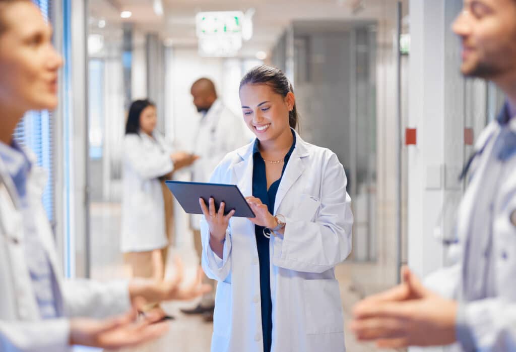 A doctor smiling and looking at her tablet in a hallway full of doctors talking.