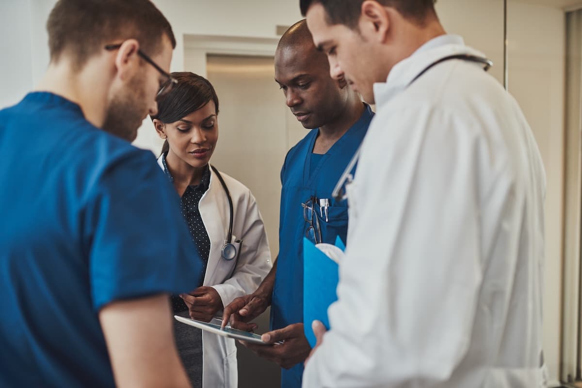 A group of multiracial doctors standing grouped closely together looking at a handheld tablet in their PCMH.
