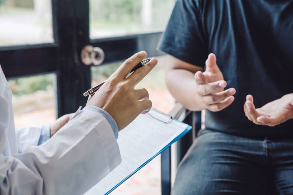 A doctor gesturing with a pen, holding a clipboard, talking to a patient.