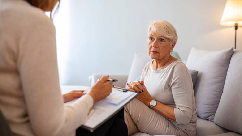 An older woman sitting on a couch, listening to her doctor go over her healthcare plan.