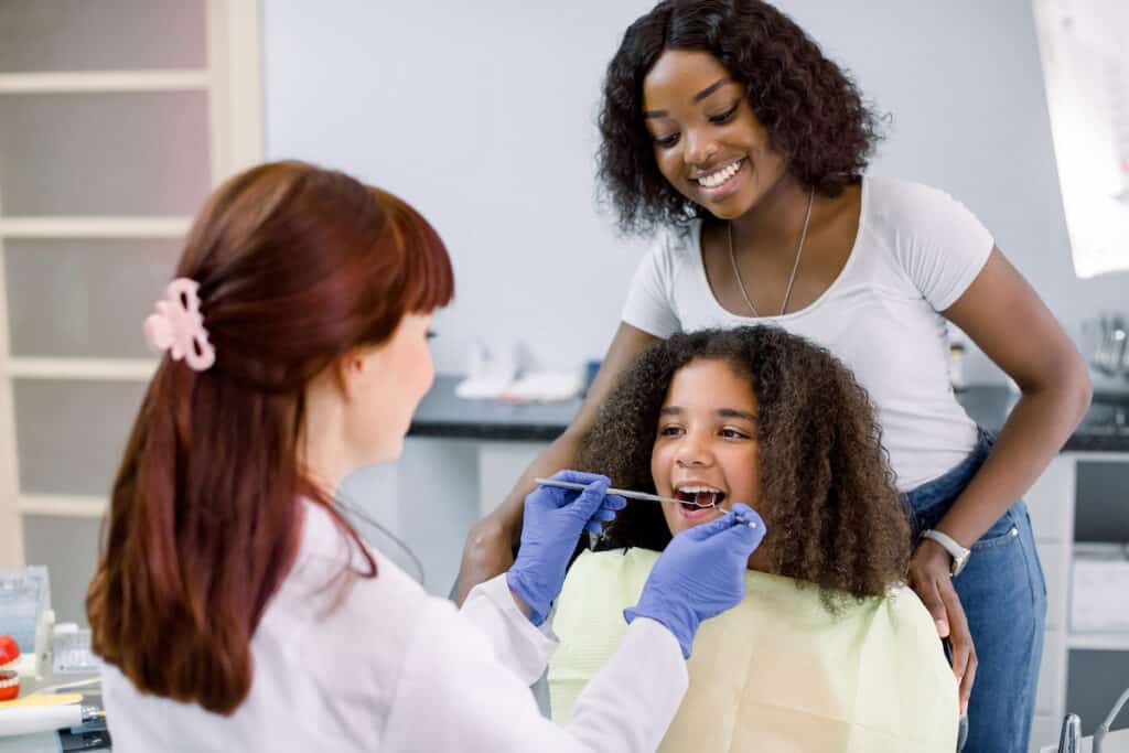 A dentist examining the teeth of a young girl with her mom standing behind her.