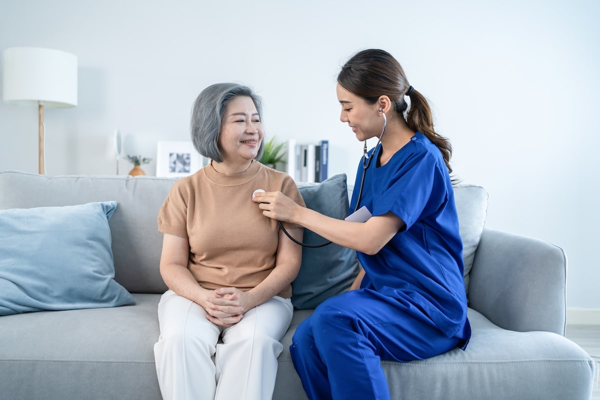 A young doctor measures heart rate with her stethoscope on her female older patient on a sofa.
